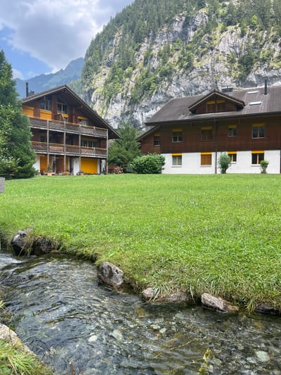 This is a photograph taken in Wengen, Switzerland, featuring a tranquil scene with natural and man-made elements. In the foreground, a clear, shallow stream flows over a bed of colorful pebbles. The stream is bordered by rocks and lush green grass that extends into the midground.
Behind the stream and grass, two multi-story wooden buildings are visible. The building on the left is a lodge-style structure with multiple balconies and yellow shutters. The building on the right is a larger, darker-roofed building with a similar wooden exterior and yellow shutters. Both buildings appear to be residential or hotel accommodations.
In the background, a steep, rocky mountainside covered in dense green trees rises dramatically. The sky above is partly cloudy with a mix of white clouds and blue sky. The overall impression is one of a peaceful mountain village setting, likely during the daytime in a season with abundant greenery. There are no people or animals visible in the image.