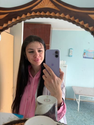 A young woman with long, dark hair takes a mirror selfie in what appears to be a room with light blue walls. She is wearing a pink hoodie and a white lab coat. She holds a lavender-colored smartphone in her right hand, capturing her reflection. The room contains a dark wood cabinet, a refrigerator, a bench with a light-colored top, and a poster on the wall. The floor is light blue with a textured pattern. A decorative wooden frame surrounds the mirror. The overall impression is that of a clinical or institutional setting, potentially a medical facility or dormitory, located in the city of Chumna, Russia.
