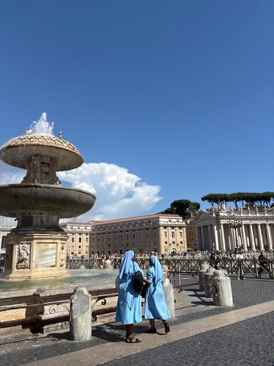 In a sunlit scene in St. Peter's Square, Vatican City, two nuns in light blue habits stand near a large, multi-tiered fountain. The fountain, constructed of stone, features water cascading from its upper basin. In the background, the colonnade of St. Peter's Basilica, adorned with statues and topped by cypress trees, is visible. Other buildings with yellow facades and red-tiled roofs are also present. The square is paved with cobblestones and demarcated by metal barriers. A crowd of tourists can be seen in the distance. The sky is a clear blue with a few white clouds.