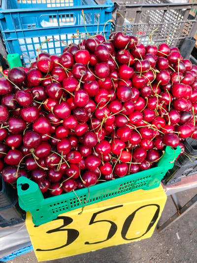 This image shows a close-up, high-angle view of a large pile of fresh cherries overflowing from a green plastic crate. The cherries are a deep red to dark purple color, with some appearing almost black. They have long, thin stems attached, and are tightly packed together.

The green crate is positioned on top of a yellow price sign, which has large, black numbers painted on it. The visible portion of the price sign reads "350". Behind the crate and sign, various stacked blue and black plastic crates are visible, suggesting a market or fruit stand setting. Some of the cherries have spilled out of the crate and onto the yellow sign.

The lighting suggests it is daytime, likely outdoors, given the bright illumination and the slight shadows. There are no people visible in the frame. The scene appears to be a vendor's display of fresh produce, likely at a market in Yessentuki, Russia. The focus is entirely on the abundance of ripe cherries.