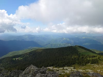 This image presents a panoramic view of a mountainous landscape, specifically located near the city of Anton, Bulgaria. The upper portion of the frame features a sky predominantly filled with large, white and light grey cumulus clouds, interspersed with areas of clear blue.
The lower two-thirds of the image depict the terrain. In the immediate foreground, dark grey and moss-covered rocks and boulders are visible, with sparse green vegetation growing between them. Immediately behind this rocky area, a dense, dark green coniferous forest occupies a significant midground slope, extending across the frame.
On the left side of the midground, a clearing within the coniferous forest reveals a cluster of buildings characterized by reddish-brown roofs and light-colored facades. To the right, the coniferous forest transitions into an exposed, grassy mountain ridge that gently rises.
In the background, multiple successive layers of mountain ranges recede into the distance, exhibiting a progressively lighter blue hue due to atmospheric perspective. These distant ranges appear covered in a mix of forested and open, treeless areas. No human figures or animal life are present in the scene. The lighting suggests natural daylight conditions.