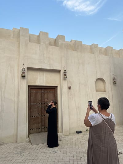 The scene depicts two adult females in an outdoor paved courtyard within a heritage district of Sharjah, United Arab Emirates. The background is dominated by a light-colored, textured wall of traditional Arabian architecture, featuring a crenellated parapet along its top edge. Centrally located in this wall is a large, recessed wooden double-door adorned with metal studs and intricate carvings. Two traditional brass and glass lanterns are mounted on the wall, one on each side of the door. To the right of the door, an arched niche is carved into the wall, with a small circular brown object mounted beneath it.
On the left side of the image, one woman, wearing a long-sleeved, floor-length black garment and glasses, stands facing slightly right. She looks upwards with her hands positioned near her face, engaged in a pose. A light-colored ramp is visible at the base of the wooden door behind her. On the right, a second woman is viewed from behind, wearing a white short-sleeved top over a long, light-brown sleeveless dress. She holds a black smartphone vertically with both hands, actively aiming it at the first woman to capture a photograph. The ground is paved with light-colored, rectangular stones arranged in a herringbone pattern. The sky above is clear blue with sparse white cloud formations. A small, dark, rectangular object rests on the paved ground near the wall, behind the second woman.