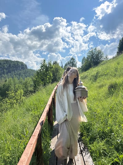 In Gubakha, Russia, a young woman with long brown hair walks along a wooden boardwalk. She is wearing a flowing cream-colored robe over a light-colored skirt and holds a miniature wooden building in her hands. She is smiling with her eyes closed, her head tilted back. Behind her, a few other people are visible on the boardwalk, walking in the same direction. The boardwalk is bordered by a wooden railing and surrounded by lush green grass and trees. In the background, a large, rocky mountain covered in trees can be seen under a bright blue sky with fluffy white clouds. The sun is shining, casting shadows on the boardwalk.