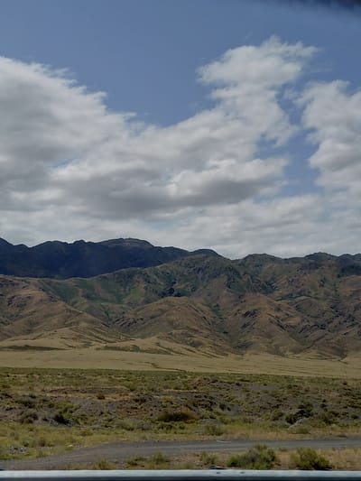 This is a photograph of a mountain range under a cloudy sky. The mountains are rugged and appear to be covered in dry vegetation, with some patches of green. In the foreground, there is a dry, scrubby landscape, and a dirt road is visible. The sky is a pale blue with scattered white and gray clouds. The image appears to have been taken during the daytime, likely in a semi-arid or arid region. There are no people or man-made structures clearly visible in the scene, aside from the road. The location is identified as being in the city of Ынталы, Kazakhstan.