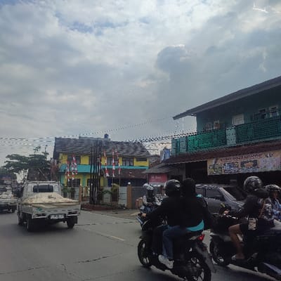 The image depicts a daytime street scene in Cimangkok, Indonesia, under an overcast sky with significant cloud cover. The paved road features a white centerline. In the foreground, multiple vehicles are observed moving away from the viewer. On the right side, three motorcycles are in motion; the foremost carries two riders wearing helmets and dark clothing, with the passenger in a light blue undershirt. Behind them, another motorcycle also carries two helmeted individuals, and a third single rider on a motorcycle is partially visible. On the left side of the road, a white pickup truck with a light-colored cover over its bed is present, bearing license plate "F 3633 CB". Another similar pickup truck is visible further down the road.

Along the right side of the road, buildings line the street. A prominent two-story building on the left features a yellow upper facade and a light blue lower facade with a dark roof. Multiple red and white Indonesian flags are displayed on poles in front of this building, behind a grey fence. To its right, another building with a dark teal or green upper section and a darker roof houses a commercial establishment on its lower level, identified by a sign partially reading "TOKO CIMANGKOK LUU". Overhead, numerous power or utility lines are visible, stretching across the frame.