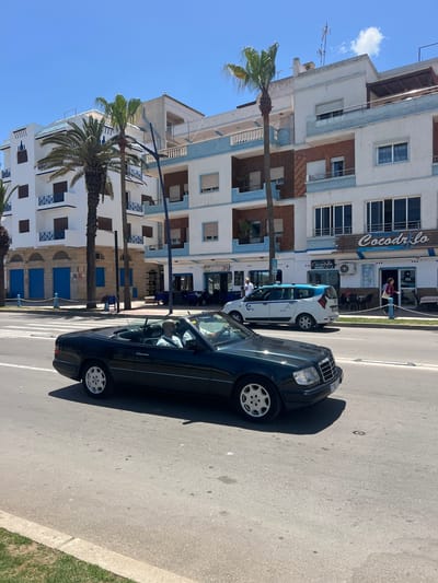 The image depicts a street scene in M'Diq, Morocco. A black convertible car is parked on the right side of the street, occupying a significant portion of the frame. The car is facing away from the viewer, and the driver is visible inside.
Buildings line the left side of the street. These are multi-story structures, mostly white with blue trim and brown brick accents. The architectural style is Mediterranean. Palm trees are planted along the sidewalk, adding to the coastal ambiance. A restaurant named "Cocodr'ilo" is visible on the building at the top of the image. A white van is parked in front of it. A person is standing in front of the restaurant. The sky is blue and clear, indicating a sunny day. The street is paved, and there is a painted white line separating the traffic lanes. A grassy area is on the extreme right edge of the frame.