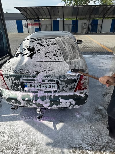 A car wash station is depicted, featuring a green sedan covered in soap foam, positioned beneath a covered area with a dark, translucent roof supported by metal poles. The car's license plate displays "HH 7153 AH". A person is actively washing the car with a brush. In the background, a striped fence separates the car wash area from a building. A sign in Cyrillic text, "ПЛАНГІ ДЛЯ БУДІВЕЛЬНИХ СИСТЕМ", is visible on the fence, along with contact information including a phone number. The ground is wet and covered in soap suds. The scene is set in the city of Chemerivtsi, Ukraine.