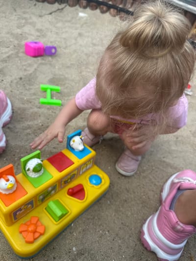 The image depicts a young child, likely a toddler, in a crouched position, playing with a yellow and blue pop-up toy featuring farm animals. The child has blonde hair styled in a bun and is wearing a pink t-shirt and pink shoes. Also visible are two pink and white sneakers, one on the child's left foot and another belonging to another person on the right side of the frame. Scattered on the sand-colored ground are a few other toys: a bright pink block and a smaller purple piece, and a green "H" shaped toy. The environment appears to be an outdoor area, possibly a backyard or playground, with sand or a similar granular surface. There is no visual information to place the scene in Donchovci, Bulgaria.