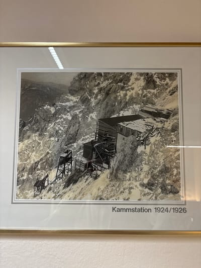 The photograph depicts a mountainous scene featuring a construction site, possibly for a cable car station, labeled "Kammstation 1924/1926". The structures are situated on a steep, rocky slope covered in patches of snow. Several wooden and metal scaffolding elements are visible, suggesting ongoing or recent construction. In the foreground and mid-ground, there are exposed rock faces and debris. The background shows a vast, rugged mountain landscape under a gray sky. The overall impression is one of a challenging, high-altitude construction project. The location is identified as being in Ehrwald, Austria.