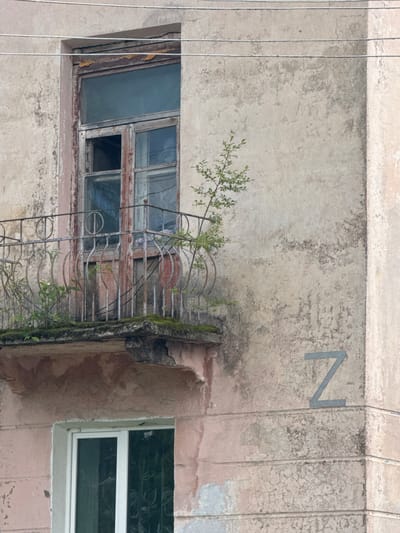This media file captures a detail of a weathered building's exterior in Teployozersk, Russia. The primary subjects are architectural elements and a small plant.
Dominating the image is a portion of a building wall, rendered in a pale, textured material that shows signs of age and wear, including cracking and discoloration. To the left, a large, old window is visible, featuring a wooden frame with peeling paint. The window panes are varied in size and some are obscured or broken, revealing a dark interior. Attached to the wall above this window is a rusty metal grate or balcony railing, with a small green plant growing through it. This plant's delicate branches and leaves are a stark contrast to the decaying structure.
To the right of the window, another window is visible, this one newer with a clean white frame and tinted glass. The contrast between the old and new windows highlights the passage of time and potential renovations or updates to the building.
In the upper right quadrant of the image, a metal letter "N" is affixed to the wall. The building appears to be an apartment block or similar multi-story structure, with power lines running horizontally across the upper portion of the frame.
The scene suggests an outdoor setting, likely during daylight hours. The overall impression is one of urban decay and the resilience of nature, as evidenced by the plant thriving on the aged facade. There are no people visible, and the time of day and weather are not definitively indicated, though the lighting suggests it is not nighttime. The location cues are the building's style, the presence of power lines, and the specific context of Teployozersk, Russia.