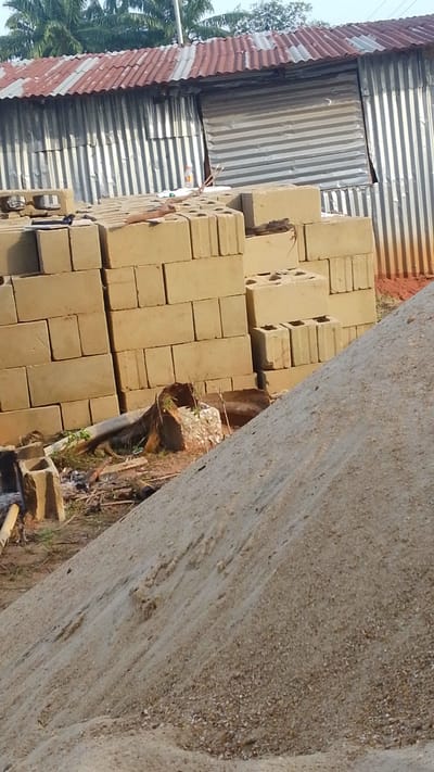 The image shows a construction site in Enugwu-Ukwu, Nigeria. A large pile of sand dominates the foreground, partially obscuring the view of stacked concrete blocks behind it. The blocks are arranged in columns, some with visible hollow cores. In the background, a corrugated metal structure with a red-roofed section is visible, indicating a temporary or informal building. Some vegetation is present.