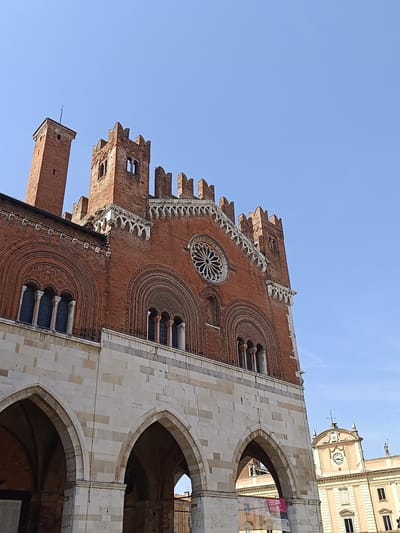 The image captures a low-angle perspective of the Palazzo Gotico in Piacenza, Italy, set against a clear, light blue sky. The building's architecture prominently features a lower level constructed from light-colored stone arches. The upper portion is made of red brick and adorned with arched windows, intricate detailing, and battlements along the roofline. To the left of the main building is a brick tower structure. In the background, the upper section of another building with a clock face and a weather vane is visible. A sign can be seen on the bottom right side of the image.