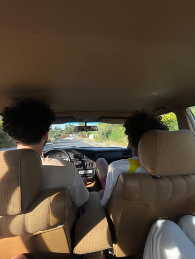 The interior of a vintage car is shown, with two people present. One person is behind the steering wheel, apparently driving, while another individual sits in the back passenger seat. The car is situated on a road, and greenery is visible through the windows, suggesting an outdoor location. Based on the metadata, the location is El Puerto de Santa María, Spain. Details like the car's dashboard, seats, and the clothing of the occupants are also visible.