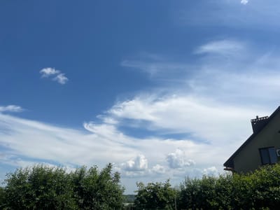 The image depicts a bright, sunny day in Koroviia, Ukraine, with a clear blue sky filled with scattered white clouds. The clouds range from wispy cirrus formations to more voluminous cumulus clouds. In the foreground, lush green trees form a dense hedge across the bottom of the frame. To the right, the angled roofline and upper portion of a multi-story house are visible. The house is painted a muted green color and features a dark roof with a chimney. A window and an air conditioning unit are discernible on the visible facade. The overall scene conveys a peaceful, rural atmosphere.