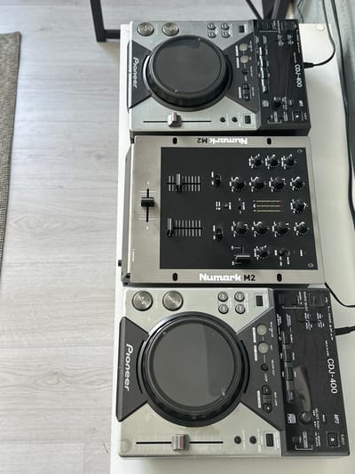 The image shows a DJ setup consisting of two Pioneer CDJ-400 CD players and a Numark M2 mixer. The equipment is arranged on a white surface, possibly a table or shelf. The CD players are positioned on either side of the mixer. The Numark M2 mixer is centrally located between the two CDJ-400 players.

The left CDJ-400 player displays a control panel with buttons, knobs, and a jog wheel. The front panel prominently features the "Pioneer" logo.

The Numark M2 mixer has multiple knobs, faders, and switches. The brand "Numark M2" is visible on the side of the mixer.

The right CDJ-400 player also shows a similar control panel to the left, with the "Pioneer" logo displayed on the front panel. Cables are visible connecting the equipment.

The background is blurred, showing a light-colored floor or surface. The image is taken from a slightly elevated perspective.

There is no location information in the image. No people, subjects, or actions are shown in the image. The objects are music DJ equipment. The environment is an interior surface, possibly a home or studio.