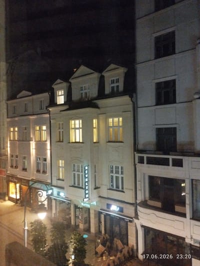 The image captures a street scene at night in Karlovy Vary, Czechia. The view is from an elevated position, looking down onto a row of buildings. The buildings are a mix of architectural styles, with some featuring ornate facades and others appearing more modern. Several businesses are visible at street level, including a hotel, cafes, and a establishment labeled "REVELTON SUITES". Lights are on in many of the windows, suggesting that the buildings are occupied. A streetlamp illuminates the foreground, casting a warm glow on the cobblestone street and some surrounding trees. The overall atmosphere is one of a quiet evening in a European city. The date and time stamp "17.06.2026 23:20" is visible in the lower right corner of the image. There are no people clearly visible in the image.