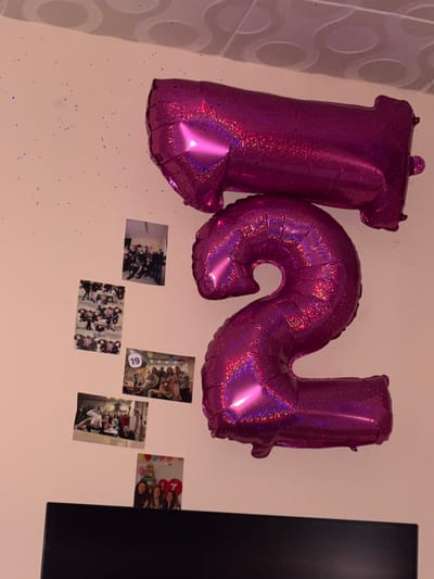 This image displays a wall decorated with two large, shiny, magenta number balloons, forming the shape of "12." Scattered on the wall are several small photographs. To the left of the balloons, there is a photograph of a group of people posing indoors. Below that, a cluster of smaller photos depicts individuals, some holding balloons. Further down, another photograph shows a group of people with a cake, marked with the number "19." To the right of this, a photograph shows a group of young women in front of a cake with the number "17" visible. The balloons and photographs are affixed to a light pink wall. Above the balloons, the ceiling has a textured pattern with circular elements. The bottom of the image shows the top of a black, flat-screen television. The scene appears to be an indoor party decoration, possibly for a birthday. The location is not specified within the image, and no information is available to determine if it is in Roza, Bulgaria.