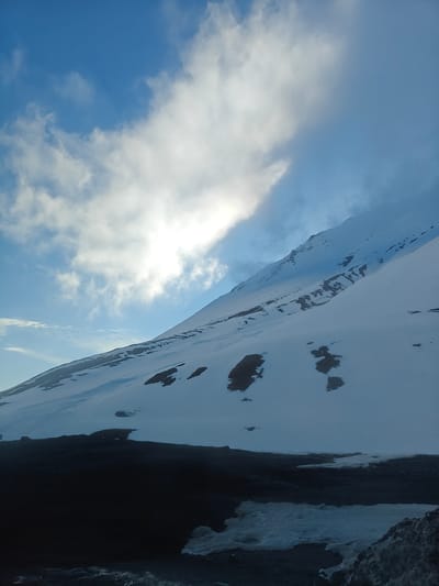 The image shows a snowy mountain landscape with a cloudy sky. The mountain slopes downward from the upper right to the lower left of the frame, covered primarily in snow with patches of dark rock or soil visible. The base of the mountain is obscured by dark shadows. The sky features a mix of blue sky and white clouds.