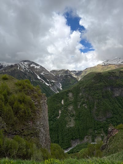The image depicts a dramatic mountain landscape with a cloudy sky. In the foreground, a steep, rocky cliff face covered in green vegetation, possibly moss and small trees, dominates the left side of the frame. The vegetation is lush and vibrant, suggesting a temperate climate.
Beyond the cliff, the scene opens up to a vast valley. The valley floor is densely forested with numerous green trees, creating a textured carpet of foliage. A narrow, winding river or stream is visible in the lower depths of the valley, reflecting the overcast sky.
Further in the distance, towering mountains rise, their slopes partially covered in patches of snow, indicating a high altitude. Some of these mountains exhibit rugged terrain with steep inclines and rocky outcrops. The sky is filled with a mix of heavy grey clouds and patches of bright blue sky, with sunlight intermittently breaking through, casting highlights on the mountain peaks.
The overall impression is one of natural grandeur and rugged beauty. The location is identified as Gudauri, Georgia, a popular ski resort town known for its stunning mountain scenery.