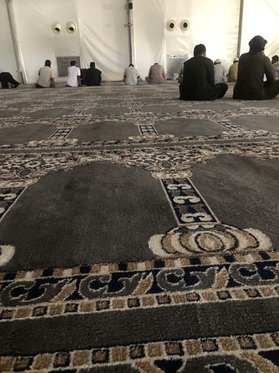 The scene depicts an interior view of a temporary prayer hall or mosque located in the city of النعيلة, Saudi Arabia. Approximately ten adult males are present, seated on a patterned grey prayer carpet. One individual on the far left is observed prostrating, while the other nine men are seated on the floor with their backs to the camera, in postures consistent with prayer or post-prayer contemplation. Their attire includes traditional garments such as white, brown, and black thobes, with some individuals wearing head coverings. The floor is covered by a grey carpet featuring intricate patterns, including mihrab arches and geometric motifs in shades of beige, brown, dark blue, and hints of red. The background consists of a white, fabric-like wall, indicative of a large tent or temporary structure. Two circular ventilation or air conditioning units are visible on this back wall, along with a partially obscured sign bearing "AL BAGHDADI" in English and Arabic text. The lighting is bright and evenly distributed throughout the space.