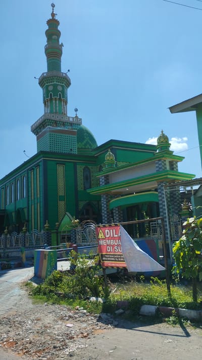 The image captures a bright, sunny day with a clear blue sky over a mosque in Cangkringsari, Indonesia. The mosque is predominantly green with intricate gold and dark green decorative elements. It features a tall, slender minaret with multiple tiers and decorative patterns. The main building has a large green dome and an entrance area with a green and gold awning supported by pillars. A fence with decorative metalwork surrounds the front of the property. In the foreground, there is a partially visible red and yellow sign with Indonesian text, stating "ADILA MEMBUA DI SU" and "PERATURAN DAERAH SID DAN MAKSIMAL RP. 50.000,- KECAMATAN SUKOD." A white flag is also visible behind the sign. The ground in front of the mosque is partially paved and partially covered in loose gravel and debris. Green foliage and bushes are present along the fence line and in front of the sign. The overall scene depicts the exterior of a religious building in a tropical setting.