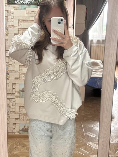 A young woman is standing in front of a mirror in what appears to be a bedroom. She is wearing a white sweatshirt with textured fringe detailing and light-wash jeans adorned with rhinestones. She is holding a white smartphone in her right hand, taking a mirror selfie. Her left hand is resting on her head, and she is looking at her reflection. The background includes a wall with patterned wallpaper and a closet or cabinet. A bed with blue and white bedding is visible in the reflection of the mirror. The flooring is a patterned wood laminate. The scene suggests a personal moment captured in an indoor domestic setting.