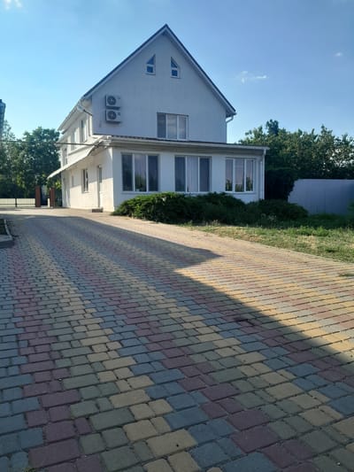 The image shows a two-story white building with an attic, situated in a paved driveway. The building has several windows, including two large ones on the first floor and several smaller ones on the second floor and attic. Two air conditioning units are mounted on the exterior wall of the second floor. The driveway is paved with multi-colored rectangular bricks. Green trees and bushes surround the building, and a metal fence is visible in the background. The sky is clear with some scattered clouds. The location is the city of Illinka, Ukraine.