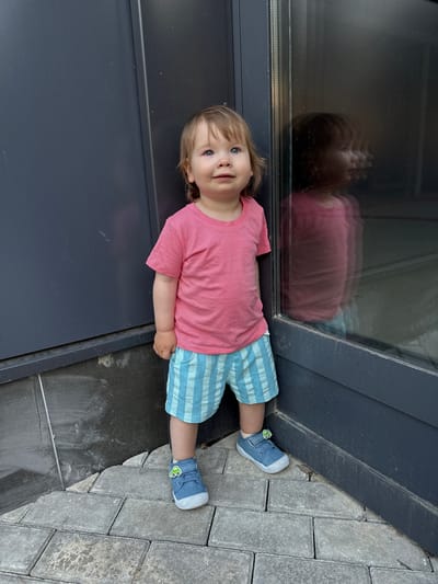 A young Caucasian child stands near a building in Kraskovo, Russia. The child has fair hair and blue eyes, and is wearing a pink t-shirt and blue striped shorts. The child's feet are clad in blue sneakers with a green car emblem on the left shoe. The child is positioned in the corner where two dark grey walls meet, with one wall featuring a reflective surface. The ground is paved with grey interlocking bricks.