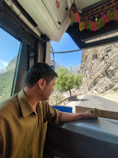 A man in a light brown shirt is seated inside a bus, looking out the window. He has dark hair and is wearing glasses on top of his head. In his right hand, he holds a green fruit, possibly an apple or pear, resting it on a surface in front of him. Outside the bus, a winding road is visible alongside a steep, rocky mountain face. A tree stands beside the road, and in the distance, snow-capped mountains can be seen against a clear blue sky. Decorative tassels, in pink and green, hang from the bus's interior ceiling. The image appears to be taken in or around Gondhla, India, given the presence of mountainous terrain and a road.