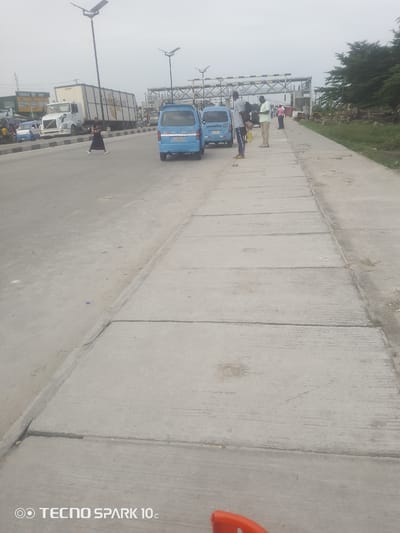 This image depicts a street scene in Eputu Town, Nigeria. The foreground features a concrete pedestrian walkway, divided into sections by visible seams. The walkway runs parallel to a paved road where several vehicles are present. These include two light blue minibuses, a white semi-truck with a large cargo container, and another smaller blue vehicle.

Several people are visible in the scene. A woman in a dark dress is crossing the road, while further down the road, three men are standing on the pedestrian walkway, seemingly waiting or observing. One man in a white shirt and beige trousers holds a yellow bag. In the background, a large metal structure, possibly an overhead pedestrian bridge or part of an ongoing construction project, spans across the road.

The weather appears overcast, suggesting either early morning or late afternoon, or a generally cloudy day. The setting is clearly an urban or semi-urban environment with significant road infrastructure. The presence of multiple vehicles, a commercial truck, and a pedestrian walkway indicates a busy thoroughfare.

The bottom of the image shows the text "TECNO SPARK 10c" with a logo, indicating it is a watermark from the device used to capture the photograph. There are no other discernible texts in the image. The overall mood is neutral, capturing a moment in the daily life of a Nigerian town.