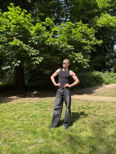 A young man stands on a grassy path in a park, likely Christchurch Park in Ipswich, United Kingdom. He is wearing a black tank top, dark jeans, and sunglasses. He is posing with his hands on his hips, flexing his arm muscles. The path is bordered by a large tree on the left and grass on the right. The tree's canopy extends overhead, creating a shaded area. The background is a mix of trees and greenery.