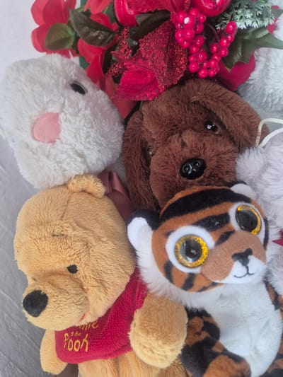 This is an overhead, close-up photograph showcasing a cheerful collection of plush toys and festive decorations, likely arranged indoors.
The central subject is a small stuffed tiger with large, prominent golden glitter eyes, a small black nose, and striped orange, black, and white fur. To its left, a brown plush animal, possibly a dog or bear, is visible with dark, button-like eyes. Partially visible above these is a fluffy white plush object.
In the bottom left corner, a white, fluffy plush toy, resembling a rabbit or lamb, is seen with small black eyes and a soft pink nose or snout. To its right, a tan-colored plush bear is identifiable as Winnie the Pooh due to its classic appearance and a red shirt bearing yellow embroidered text that reads "Winnie the Pooh."
Intertwined with the plush toys, especially on the left side of the frame, are elements of a holiday or festive decoration. These include clusters of shiny red berries, green artificial foliage such as pine needles and leaves, and various red fabric elements, including glittery ribbons and red flower petals.
The items appear to be small, suggesting they could be ornaments, keychains, or part of a decorative arrangement. The overall atmosphere conveyed is one of warmth and festivity. The specific location context for this media file is Krumovo, Bulgaria. No time of day or outdoor weather conditions are discernible from the image.