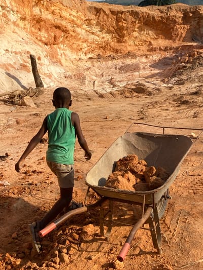 A young person, presumably a child, stands next to a wheelbarrow filled with red-brown rocks or ore. The child wears a green tank top and light-colored shorts. The environment appears to be a mine or quarry with a tall, sandy cliff face in the background. The ground is dirt, scattered with rocks and tire tracks. A wooden log or root lies nearby. The entire scene conveys an impression of hard labor in a harsh environment.