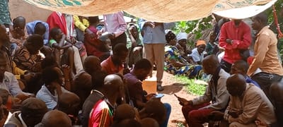 A large group of approximately 30-40 individuals is gathered outdoors in Kundu 1, Democratic Republic of the Congo, under a makeshift canopy constructed from light-colored fabric, possibly repurposed sacks, to provide shade. The majority of the attendees are adults, with a mix of males and females, and a few younger individuals also present. Most are seated directly on the reddish-brown dirt ground, some with visible green vegetation patches, while a few stand. Attire consists of varied casual clothing, including collared shirts, t-shirts, dresses, and headwraps. Several individuals are engaged with small electronic devices, likely mobile phones, which they hold and view in their hands (e.g., in the foreground and center). A man in a pink collared shirt standing center-left holds a notebook and pen, seemingly taking notes. The overall demeanor of the crowd appears attentive and engaged. Dense green foliage is visible in the background, indicating an outdoor, vegetated setting. The scene is brightly lit by natural light, casting soft shadows.