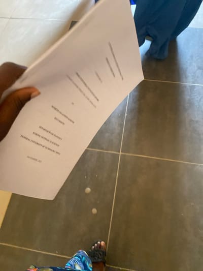 A hand holds a document displaying the title "SIMULTANEOUS DETERMINATION OF SPECTROPHOTOMETRICALLY" with the author listed as "ROQEEB SALAUDEEN KHOIME." It indicates the "DEPARTMENT OF PHYSICS," "SCHOOL OF PHYSICAL SCIENCES," and "FEDERAL UNIVERSITY OF TECHNOLOGY MINNA." The date on the document is "NOVEMBER, 2021." The background includes a dark-tiled floor, a portion of a dark blue garment, and a foot clad in a sandal.
