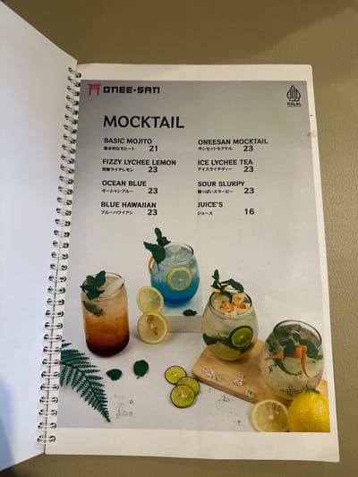 An open menu book is displayed, showcasing a "MOCKTAIL" section with several beverage options. The page is spiral-bound on the left side. A "HALAL" logo is visible at the top left corner of the page. The menu lists various mocktails with their names in English and Japanese, along with corresponding prices.
Six distinct mocktails are visually presented in different glasses, garnished with citrus slices (lemon and lime) and mint leaves. Ice cubes are scattered around the glasses, some resting on a small wooden board. One drink appears blue ("OCEAN BLUE" or "BLUE HAWAIIAN"), while others are clear, light brown, or feature fruit slices like lychee.
Visible text includes:
- "ONEESAN" (logo)
- "HALAL"
- "MOCKTAIL"
- "BASIC MOJITO" (21), with Japanese "基本的なモヒート"
- "FIZZY LYCHEE LEMON" (23), with Japanese "炭酸ライチレモン"
- "OCEAN BLUE" (23), with Japanese "オーシャンブルー"
- "BLUE HAWAIIAN" (23), with Japanese "ブルーハワイアン"
- "ONEESAN MOCKTAIL" (23), with Japanese "サンセットモクテル"
- "ICE LYCHEE TEA" (23), with Japanese "アイスライチティー"
- "SOUR SLURPY" (23), with Japanese "酸っぱいスラービー"
- "JUICE'S" (16), with Japanese "ジュース"
The prices range from 16 to 23, likely indicating a local currency. The image captures the menu from a slightly overhead perspective, highlighting the drinks and their descriptions.