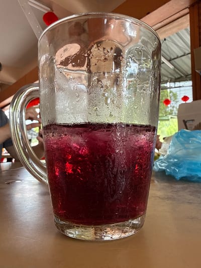 The image shows a glass mug filled with a clear liquid on the left and a dark red liquid on the right. The mug has condensation on its exterior. It is placed on a tan-colored table. The handle of the mug is silver. In the background, there is a roof structure with clear roofing panels and two red lanterns. A person and blue plastic are visible blurred in the background. It appears the photo was taken indoors, near an opening to the outside. The location is George Town, Malaysia, inferred from the style of lanterns.