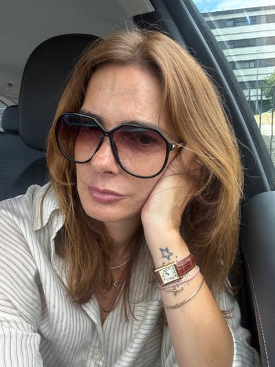 This is a close-up, low-angle selfie of a woman inside a car. The woman has long, light brown hair and is wearing oversized black sunglasses. She is resting her head on her hand, and her left arm is visible, adorned with a watch and several bracelets, including one with a small cross charm. She also has a small star tattoo on her wrist. She is wearing a light-colored, pinstriped button-up shirt. The background of the image shows a building with vertical black and white stripes, possibly seen through the car window. The lighting suggests it is daytime. The overall mood is casual and relaxed. The image is geotagged to Loures, Portugal.