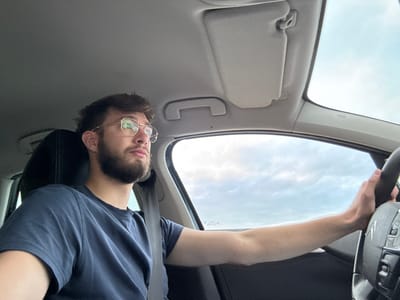 A male individual is observed from the mid-chest up, seated in the front driver's position of a vehicle in Châtenois, France. He possesses short brown hair, a beard, and wears gold-rimmed hexagonal glasses. His attire consists of a dark blue short-sleeved t-shirt, and a black seatbelt is correctly positioned across his right shoulder and chest. His right hand is actively gripping a black multi-function steering wheel, featuring visible control buttons on the right spoke and a central emblem resembling a 'C' logo. The individual's head is turned slightly to the right, and his gaze is directed forward and slightly upward, beyond the vehicle's windshield. The vehicle's interior ceiling is light grey, with a matching sun visor in the raised position on the passenger side and an overhead grab handle. Through the vehicle's windows, a cloudy daytime sky is visible, characterized by variations in light intensity. No other individuals or external landmarks are discernable.