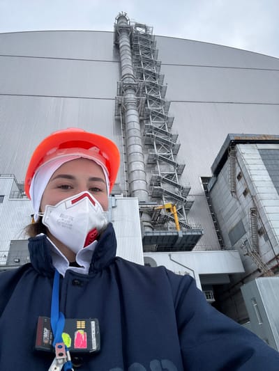 A female individual is depicted in an outdoor selfie, looking directly at the camera. She wears an orange hard hat over a white head covering and a white FFP3 respirator mask, identified as "HIGH-3K FFP3 NR D" with CE 0194 marking, covering her mouth and nose. She is dressed in a dark blue jacket with a white collar visible underneath. Two electronic devices are clipped to her jacket via a blue lanyard: a black personal dosimeter marked "MGP SMC 2000 R" and another black rectangular device with a pink/yellow sticker displaying "488091" and "NBT6714".

The background features the massive, corrugated metallic structure of the New Safe Confinement (NSC) arch, which covers Reactor 4 of the Chernobyl Nuclear Power Plant. A tall, slender industrial structure, equipped with multiple platforms and ladders, is prominently visible against the NSC, extending vertically. To the right, an ancillary concrete industrial building with visible windows or vents is present. The sky above is uniformly overcast and light grey. The scene is located at the Chernobyl Nuclear Power Plant, near the city of Pryp'yat', Ukraine.