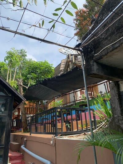 The image shows a view of a patio area with a black awning overhead, located in the city of Kecamatan Borobudur, Indonesia, at coordinates -7.606 latitude and 110.18 longitude. The patio has a black metal railing and is decorated with a vibrant blue, pink, and yellow color scheme. There are wooden tables and chairs on the patio, and a large tree growing in the background. The patio is situated beneath a concrete structure with a network of electrical wiring and overhead cables running along the top. There are green plants and foliage surrounding the patio, suggesting that the location is a restaurant or cafe. The patio is located on a slight incline, and there is a wooden staircase leading down to a lower level. The overall scene suggests an outdoor dining experience in a vibrant and colorful setting.