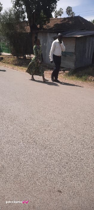 The image captures an outdoor street scene in Homa Bay, Kenya, under clear daylight. A paved asphalt road surface occupies the lower two-thirds of the frame. Two individuals are depicted walking from left to right on a dirt shoulder parallel to the road. The lead individual is a female, wearing a green patterned long-sleeved dress and light-colored flat shoes. Following her is a male subject, dressed in a light-colored long-sleeved button-down shirt and dark trousers, holding a dark-colored mobile phone to his right ear, suggesting he is engaged in a call. In the background, to the right, stands a single-story structure with weathered corrugated metal walls and a brown tiled roof. Beyond this structure, several trees, including identifiable palm trees, are visible against a light blue sky with scattered white clouds. Green foliage and a large tree trunk are present in the left background. Strong sunlight casts distinct shadows of the individuals to their right on the dirt ground.