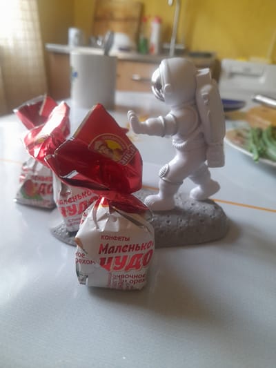 The image shows a white astronaut figurine posed on a gray, moon-like surface. The figurine is depicted in a dynamic stance, with its arms and legs positioned as if in mid-movement. In front of the astronaut, there are several candy wrappers. One wrapper is predominantly red with some yellow detailing and appears to be partially crushed. Another wrapper is white with red lettering. Visible text on the wrappers includes "КОНФЕТЫ" (candies), "Маленькое" (little), and "ЧУДО" (wonder). The background suggests an indoor setting with kitchen-like elements such as counters and cabinets, illuminated by natural light, implying it is daytime. The overall scene is casual and likely takes place in a home environment.