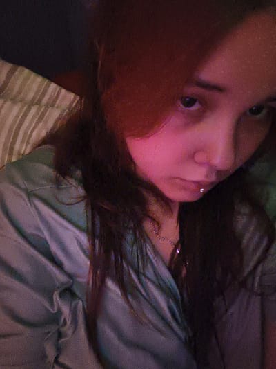 The image is a close-up, dimly lit selfie of a person with dark hair, wearing a light-colored, possibly satin, garment. The person has a piercing in their lower lip. The background appears to be a bed with striped bedding. The lighting casts a pinkish hue on the person's face. No specific location in Ekibastuz, Kazakhstan is discernible.