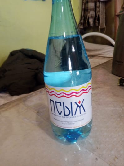 This media file is a close-up shot of a plastic bottle of sparkling water. The bottle is predominantly teal green with a clear, blue-tinted liquid inside. A white label is affixed to the lower portion of the bottle. The label features a bold, dark blue Cyrillic text that reads "ПЫХ". Below this, in smaller white text, is "АКТИВАТОР ЖИЗНЕННОЙ ЭНЕРГИИ" and below that, "ПРИРОДНАЯ ГАЗИРОВАННАЯ ВОДА С КАВКАЗА". The label is bordered by wavy lines of yellow, pink, and red at the top.

The bottle is placed on a patterned surface that appears to be a table or floor, with a light brown and beige speckled texture. In the background, to the left, is a dark brown, quilted jacket or blanket draped over something unseen. Further back, a wall is visible, painted a pale yellow. To the right of the bottle, a portion of a dark brown object, possibly an appliance like a coffee maker or kettle, is visible. A white curved object, perhaps part of a chair or bed, is also in the background. The overall lighting suggests an indoor setting, possibly during the day. The scene appears to be a casual, domestic environment. No people are visible in the frame.