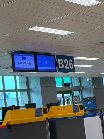 The image is taken at an airport. The gate number "B26" is prominently displayed above two monitors. The lower monitor indicates that the flight, BA 7304, to London City is delayed on arrival. In the background, windows reveal a view of an airport terminal. A yellow kiosk-like structure with advertising is to the right of the signage. The Italian cities "Milano Linate" is visible in the display of the advertising. The brand "IT/A AIRWAYS" and "15 min." also are visible in the kiosk signage. The location is near Peschiera Borromeo, Italy, as the Linate Airport is the Milanese airport closest to the city of Peschiera Borromeo.