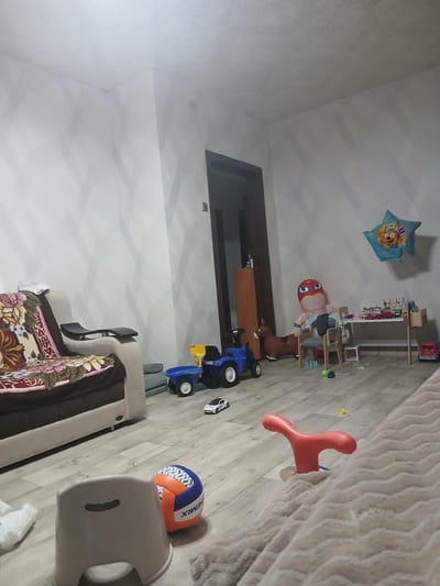 The image depicts a child's playroom within a residential dwelling located in Kastornoye, Russia. The room features light-colored walls with a geometric pattern and a light wooden floor.
Prominent objects include a beige sofa with a floral-patterned throw blanket, a small grey stool, a blue and orange volleyball, and a red ride-on toy. Scattered across the floor are various children's toys: a blue toy truck with a trailer, a brown toy horse, a large inflatable character resembling a wrestler, a white toy car, and a wooden play table with small toy vehicles and building blocks. A blue star-shaped balloon with cartoon characters is suspended from the ceiling. A wooden wardrobe stands against the wall near a doorway.
The lighting suggests the image was taken indoors, possibly during daylight hours, with artificial light sources contributing to the overall illumination. The room appears to be in a state of use, with toys distributed across the floor, indicating recent or ongoing play activity. There are no people visible in the frame. The overall impression is that of a lived-in space designed for children's entertainment and play.