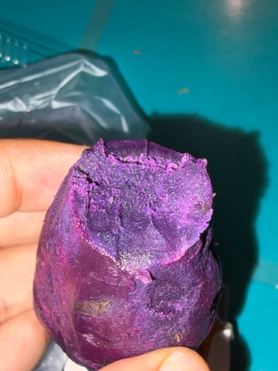 A close-up shot shows a hand holding a bright purple sweet potato. The sweet potato is roughly oval-shaped and has a textured, slightly crumbly surface. It appears to have been peeled or broken open, revealing the vibrant purple flesh. The background is a bright teal surface, possibly a table, with a dark shadow cast by the sweet potato and the hand. A portion of a clear plastic bag is visible in the upper left quadrant of the image. There is no identifiable information about the location in Tupah, Indonesia.
