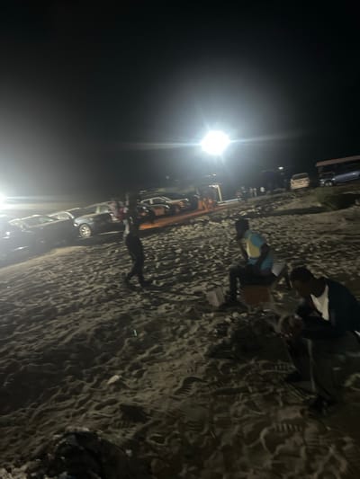 The image shows a nighttime scene in Isunba, Nigeria. Several cars are parked on what appears to be a sandy or gravely surface, possibly a beach or shoreline. A bright light source illuminates the scene, creating strong shadows. People are gathered, some standing near the vehicles and others sitting or interacting in the foreground. Details are obscured by the lighting conditions, but the presence of vehicles and people suggests a gathering or event.