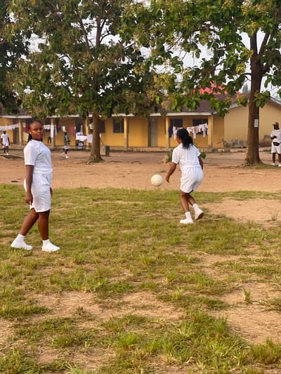 The image depicts two young women in white uniforms on what appears to be a school compound. One woman is in the foreground, facing the camera, with her left hand resting on her hip. She has dark, curly hair braided and is wearing white socks and shoes. Behind her, and to her left, is a large tree with green leaves and small, light-colored buds or fruits.

Further in the background, there is a yellow building with red-tiled roofing. Clothes are hung on a line in front of the building. The ground is a mix of dirt and dry grass. Another woman, also in a white uniform, is visible in the mid-ground, in a dynamic pose, appearing to be kicking a white ball. She is facing away from the camera and is also wearing white socks and shoes. The overall scene suggests an outdoor activity or training session. The lighting indicates it is likely daytime, possibly late afternoon, given the warm tones. There is no visible text in the image. The setting appears to be a school or training facility in Emure-Ekiti, Nigeria, as suggested by the context. The individuals seem engaged in physical activity.